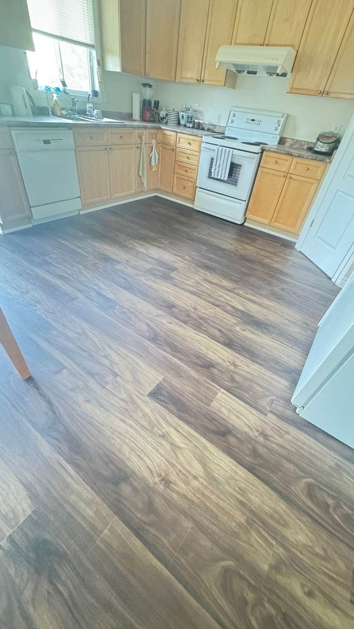 Brown wood-look laminate flooring installed in a residential kitchen with light wood cabinets, white appliances, and a window.