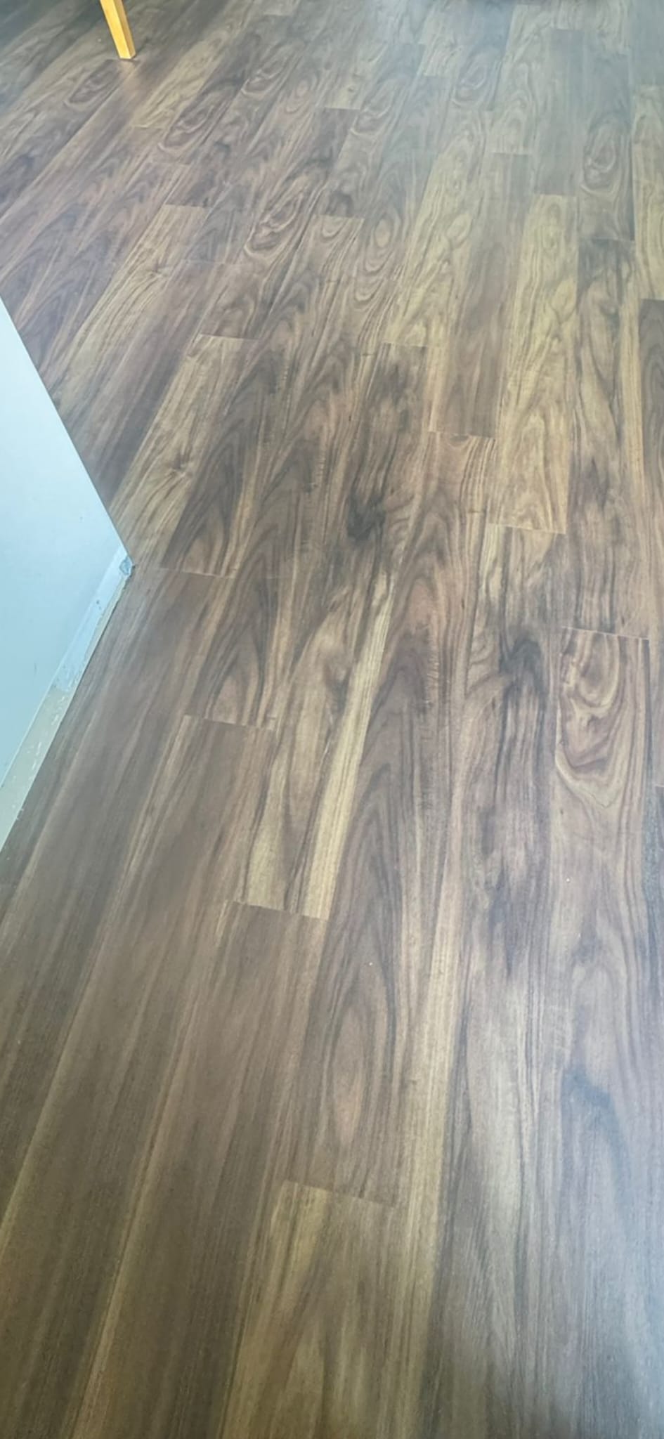 Brown wood-look vinyl plank flooring shown from an elevated perspective, with a white wall baseboard visible on the left and a small yellow leg in the top-left corner.