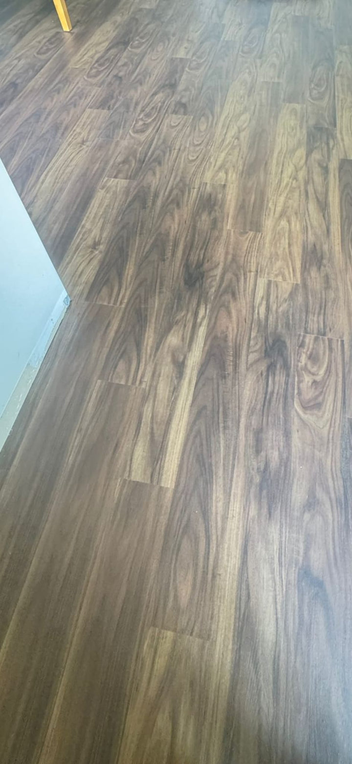 Brown wood-look vinyl plank flooring shown from an elevated perspective, with a white wall baseboard visible on the left and a small yellow leg in the top-left corner.