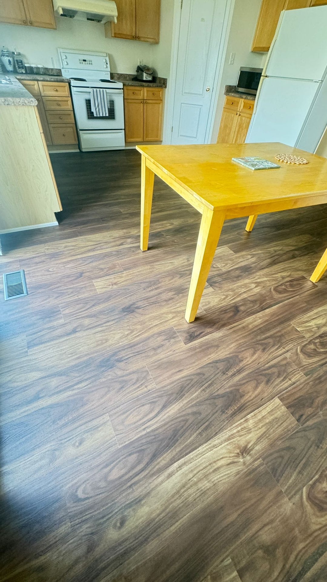 Brown wood-look vinyl plank flooring installed in a kitchen and dining area, featuring a yellow wooden dining table in the foreground.