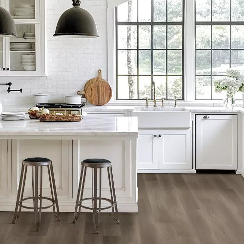 A bright, modern farmhouse kitchen featuring light brown wood plank flooring, white cabinets, a white marble island, and two dark metal bar stools.