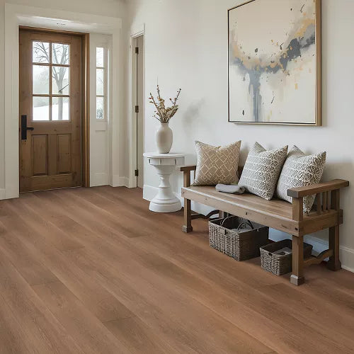 A bright, modern entryway featuring warm medium-brown hardwood flooring, a rustic wooden bench with patterned throw pillows, a white side table with a vase, and a framed abstract painting on the wall.