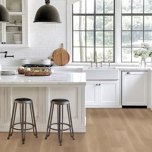 A bright, modern kitchen interior with white cabinetry, a white farmhouse sink, large black-framed windows, light wood flooring, and two dark metal bar stools at the island.