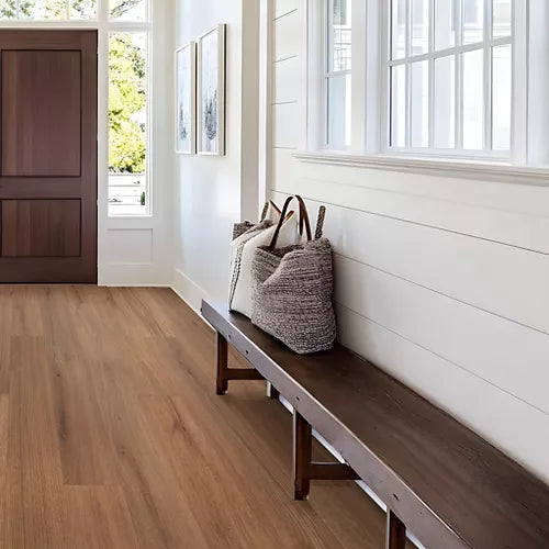 A bright entryway with light brown wood flooring, white shiplap walls, a dark wooden bench, and a woven tote bag resting on the bench.