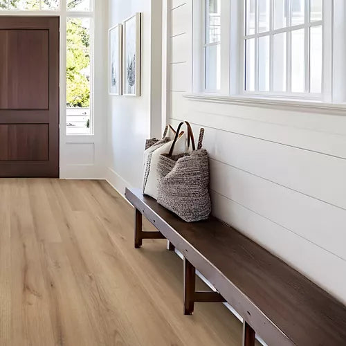A bright entryway with light oak wood flooring, white shiplap walls, a dark wood bench, and a woven straw tote bag resting on the bench.