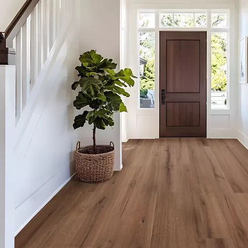 Brown wood-look flooring shown in a bright entryway with white walls, a staircase on the left, and a front door at the end of the hall. A potted green plant sits near the wall.